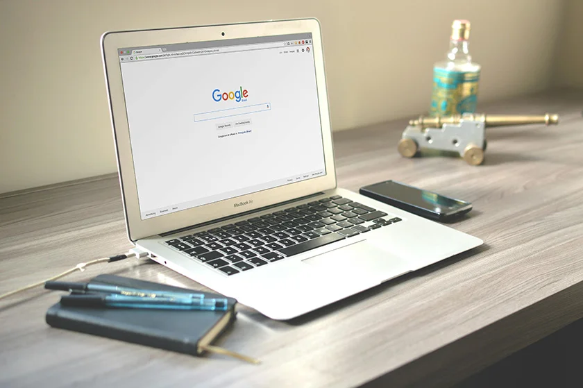 Open MacBook Air displaying the Google homepage on a wooden desk, with a smartphone, notebook, pens, and other decoration nearby