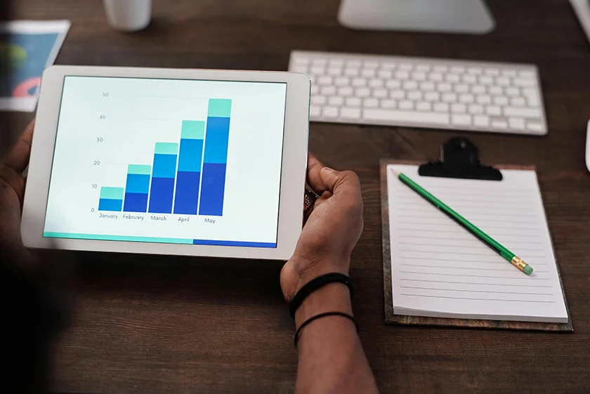 Persona sosteniendo una tablet que muestra un gráfico de barras con el crecimiento de datos mensuales de enero a mayo, junto a un portapapeles con un bloc de notas y un lápiz verde sobre un escritorio